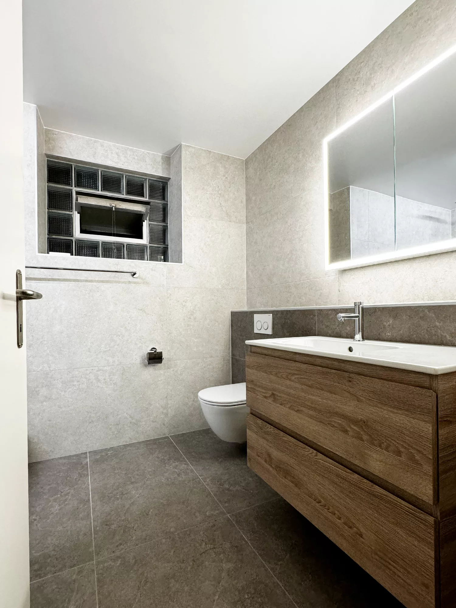 Salle de bain moderne avec meuble en bois, miroir éclairé et toilettes suspendues, carrelage gris et murs en pierre.