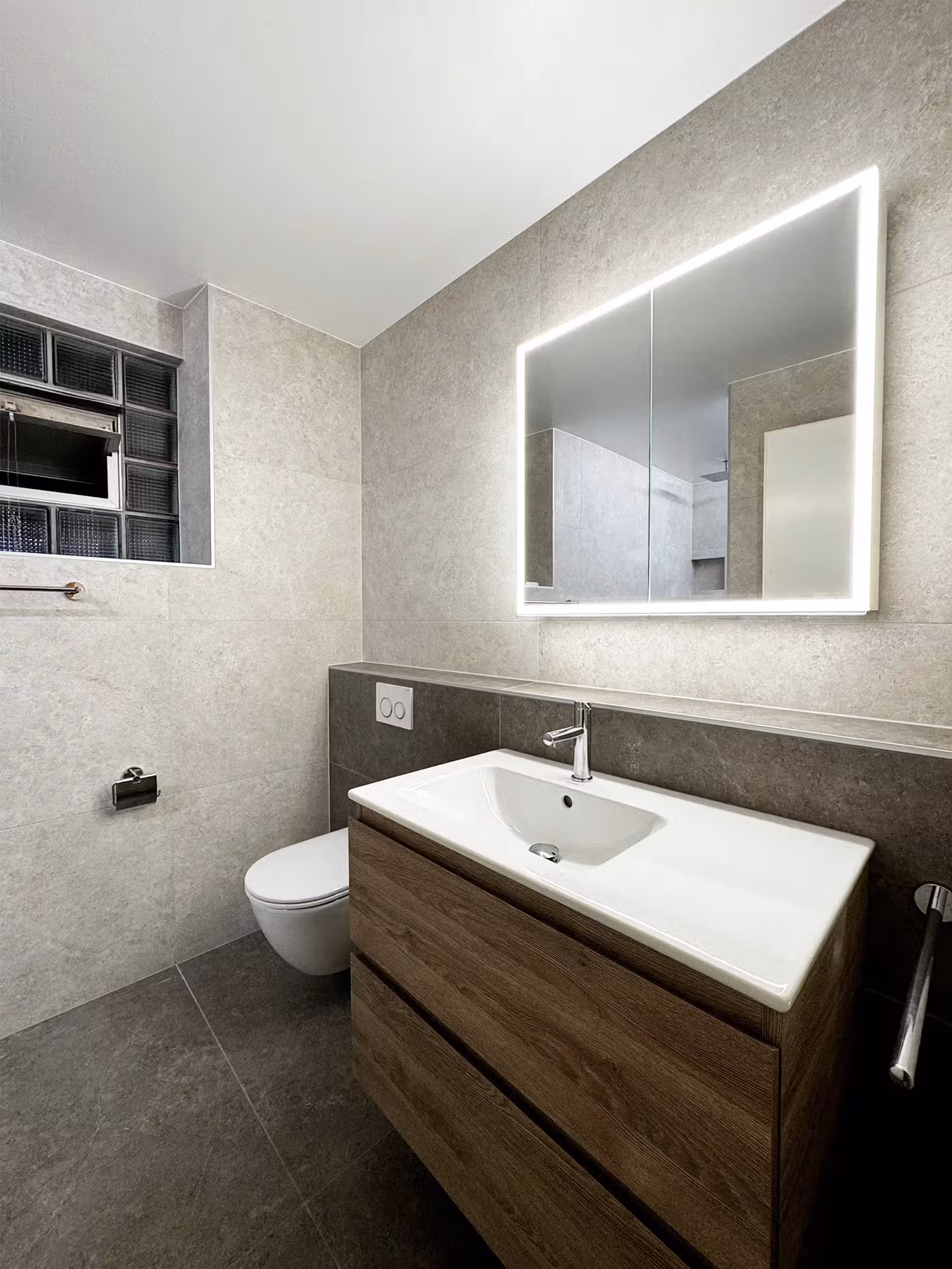 Salle de bain moderne avec meuble en bois, lavabo blanc et miroir lumineux, carrelage gris, et toilettes suspendues.