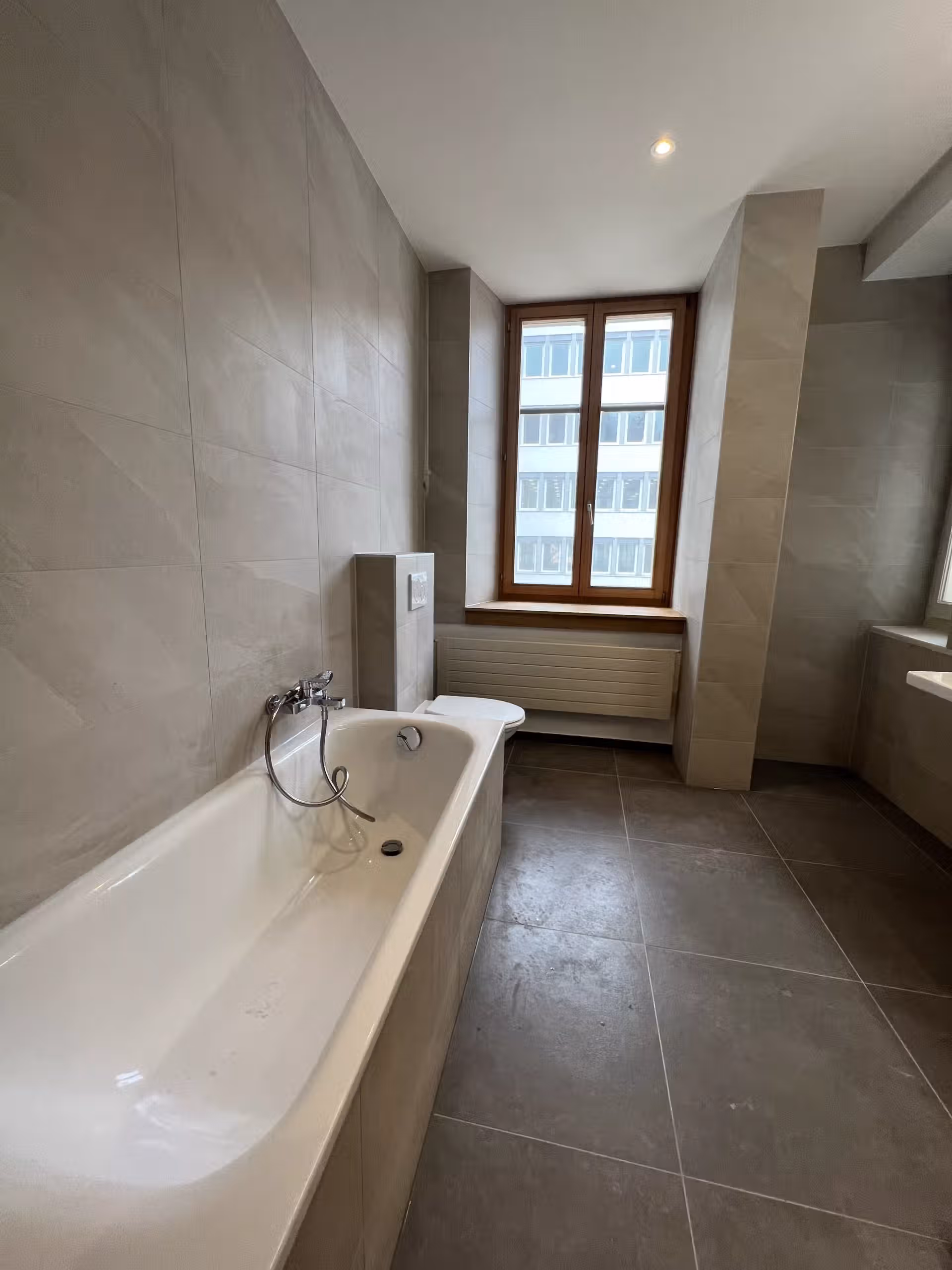 Salle de bain spacieuse avec baignoire blanche, murs en carrelage gris et fenêtre laissant entrer la lumière naturelle.